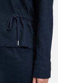 Granatowa lniana bluzka z regulowanym pasem, z długim rękawem i mankietami. Widoczna gładka faktura i subtelny połysk.