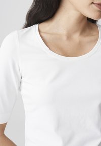 Frau mit langen dunklen Haaren, die ein einfaches weißes kurzärmliges Shirt trägt, von den Schultern bis zur Mitte des Oberkörpers vor einem grauen Hintergrund gezeigt.