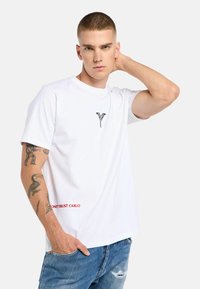 Young man with short hair wearing a white t-shirt with black logo and red text, tattooed arm, and blue distressed jeans, posing with hand behind head.