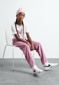 Chemise blanche à manches courtes avec un texte graphique rose, pantalon en velours côtelé rose, bonnet en tricot rose, baskets blanches avec des accents noirs, assis sur une chaise.