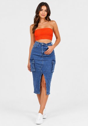 Mujer con un top corto naranja sin tirantes, falda vaquera azul de talle alto con abertura frontal y bolsillos, y zapatillas blancas caminando hacia adelante.
