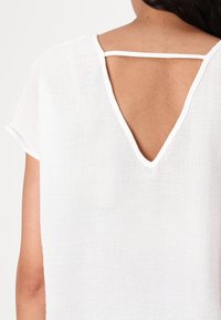Biała lniana bluzka z dekoltem w kształcie litery V z podwójnym paskiem na plecach. Krótkie rękawy i fakturowany materiał dopełniają minimalistyczny wygląd.