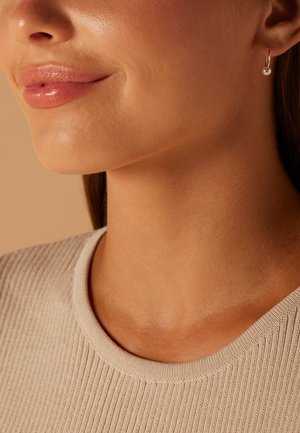 Goldene Creolen mit einem einzelnen Perlenakzent. Glatte, glänzende Textur. Modell trägt einen beigen ribbeligen Pullover mit rundem Halsausschnitt.