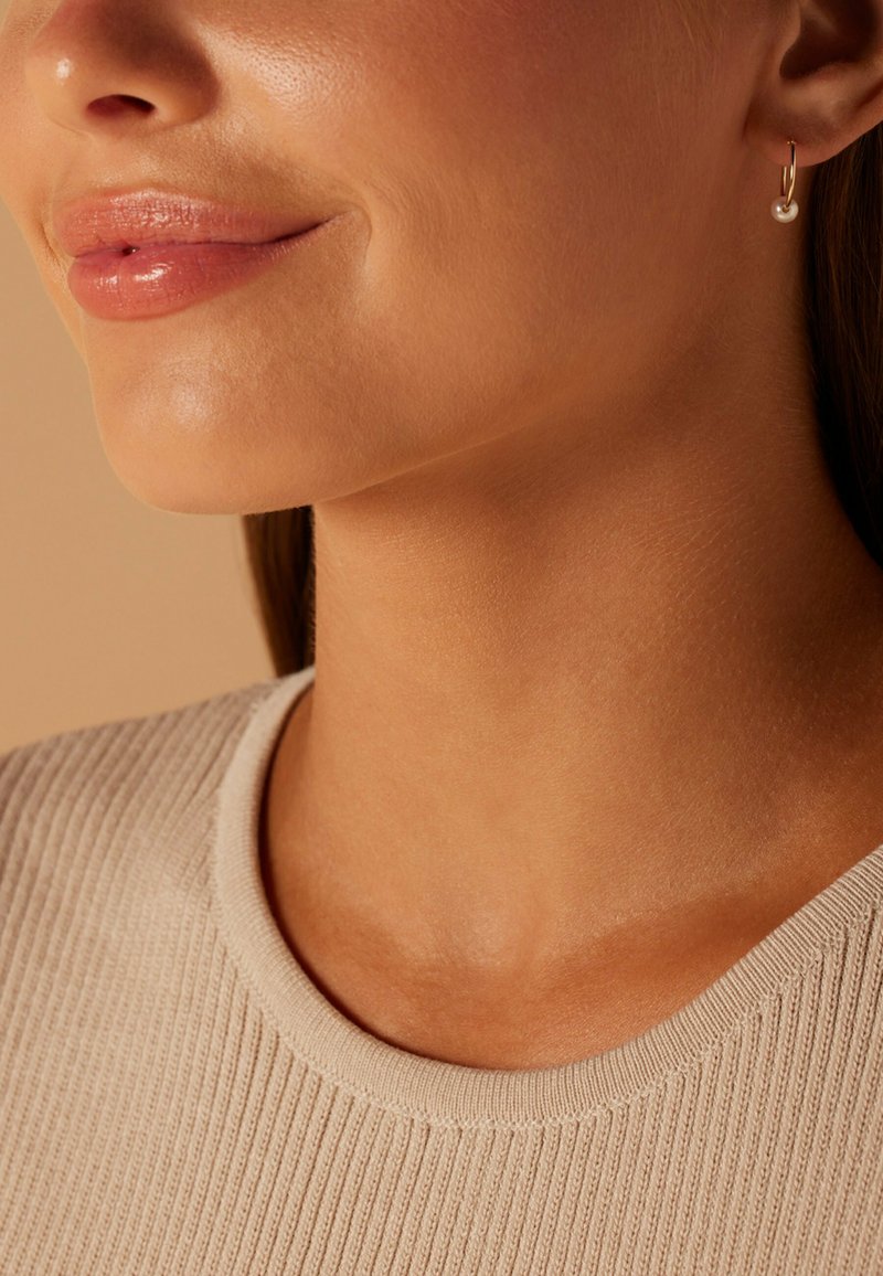 Gouden creolen met een enkele parelaccent. Glad, glanzend oppervlak. Model draagt een beige ribbeltrui met een ronde hals.