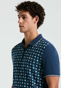 Polo shirt avec fermeture éclair, présentant un motif carré bleu et vert jade à l'avant, un dos bleu uni et un col contrastant.