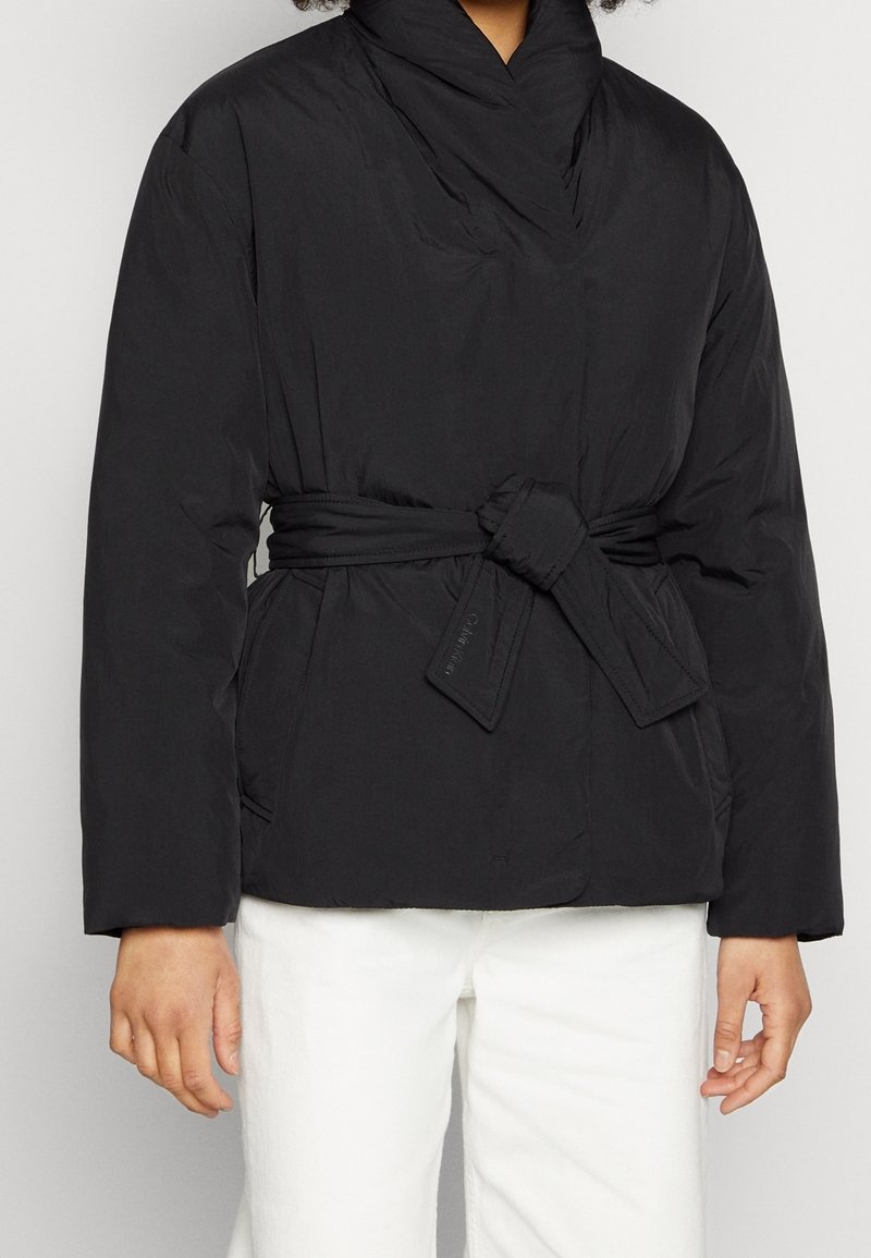 Svart puffjacka med en omslag design, med en bälte i midjan, hög krage och slät yta. Horisontell sömnad är synlig.