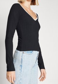 Zwarte geribbelde langemouwen top met een V-hals en witte bies, een getailleerd ontwerp, gecombineerd met lichtblauwe spijkerbroeken.