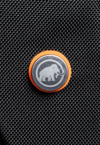 Cirkulär knapp med en metallisk silverelefantdesign på en texturerad svart bakgrund, omgiven av en orange kant.