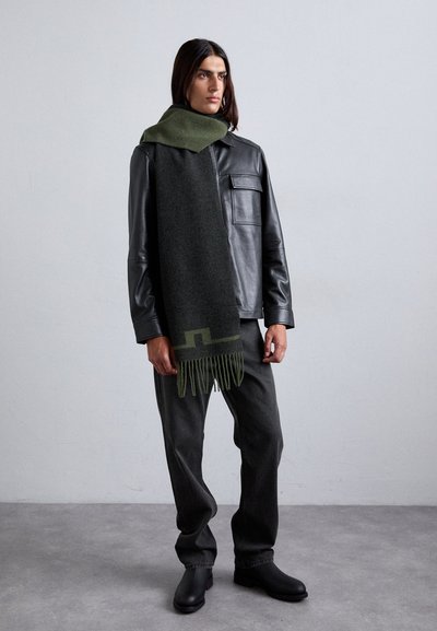 Écharpe en laine gris foncé avec des accents verts, bordures frangées, associée à une veste en cuir noir et un pantalon sombre, portée par un mannequin masculin.
