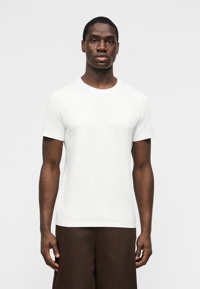 Young man wearing a plain white short-sleeve t-shirt and dark brown pants, standing against a plain light background.