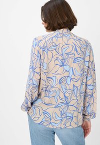 Camicetta a maniche lunghe in tessuto beige con motivi floreali blu. Texture liscia, vestibilità ampia e orlo arrotondato.