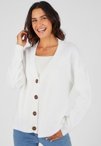 Cárdigan blanco de punto con escote en V, textura acanalada y grandes botones marrones en la parte delantera. Las mangas son ligeramente oversize.