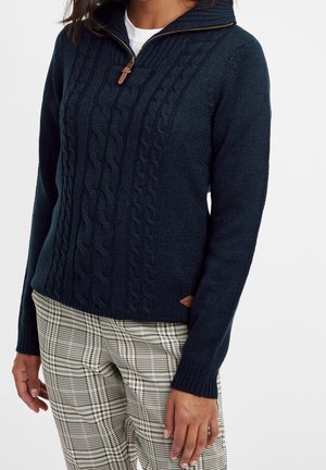 Woman wearing a navy blue cable knit sweater with half zipper and plaid beige and black pants, standing against a white background.