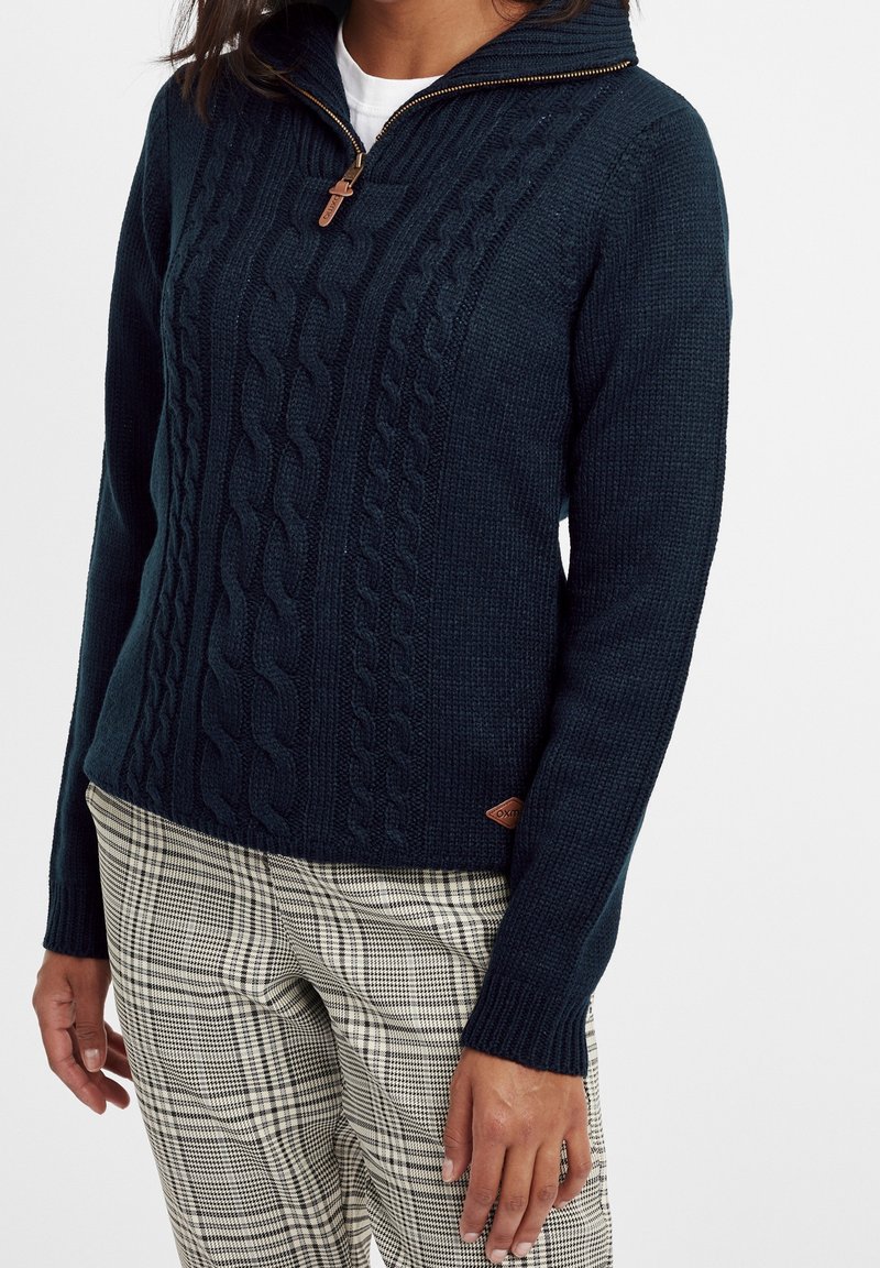 Femme portant un pull en maille torsadée bleu marine avec une demi-fermeture éclair et un pantalon à carreaux beige et noir, debout sur fond blanc.