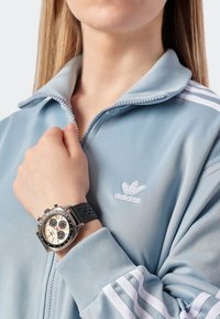 Svart klocka på en handled, med silverfärgad urtavla och tre undergrupper, bärs med en ljusblå zip-up träningsjacka med vita ränder.