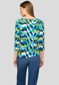 Frau von hinten, trägt ein mehrfarbiges Oberteil mit geometrischem Muster, dreiviertellangen Ärmeln und blaue Jeans vor einem schlichten Hintergrund.