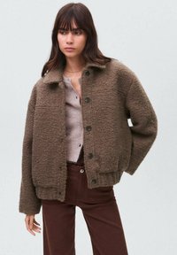 Jachetă texturată maro din material moale, cu guler, nasturi frontali și tăietură lejeră, asortată cu un pulover deschis la culoare și pantaloni maro.
