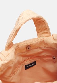 Sac fourre-tout couleur pêche avec poignées texturées, intérieur doux, poche zippée et étiquette de marque. Tissu lisse avec un accent cousu.