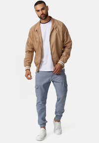 Chaqueta bomber marrón con puños acanalados, camiseta de cuello redondo blanca y pantalones cargo azul claro. El modelo lleva zapatillas blancas.