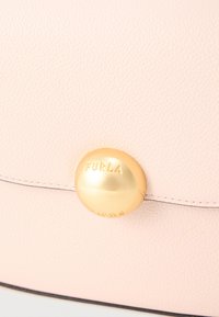 Világos rózsaszín bőrtáska, amelyen feltűnő, kerek arany kapocs található, amelyen a "Furla" felirat dombornyomott, valamint kontrasztos varrások az éleken.