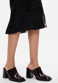 Persoană îmbrăcată într-o fustă texturată neagră și mule cu toc înalt din piele patentată maro închis, cu tocuri tip block, pe un fundal alb.