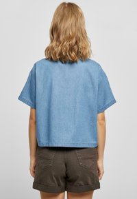 Person with shoulder-length wavy blonde hair wearing a loose light blue short-sleeve top and dark rolled-up shorts, facing away.