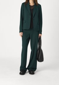 Costume vert sur mesure avec un blazer à un bouton et un pantalon à jambes larges. Porté avec un haut noir transparent et un sac noir. Des chaussures noires complètent la tenue.