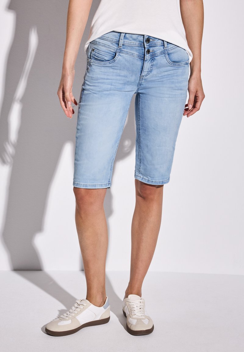 Ljusblå denimbermuda med en figursydd design, femficksstil och en lätt blekning. Matchas med vita sneakers.
