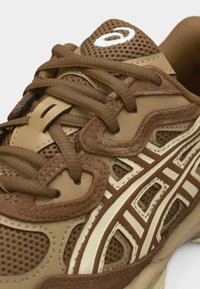 Zapatilla deportiva marrón y beige con tela de malla, superposiciones de ante y rayas estampadas, con cordones marrones atados sobre la lengüeta.