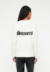 Biały, puszysty sweter z czarną, stylizowaną grafiką "Sinners" na plecach. Żebrowany dół i mankiety, luźny krój.