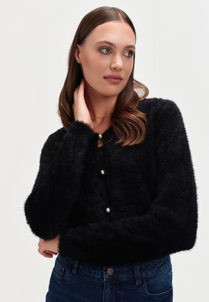 Kobieta z brązowymi włosami, ubrana w czarny puszysty kardigan z perłowymi guzikami i ciemnoniebieskie dżinsy, na jednolitym białym tle.