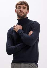 Mann mit Bart, der einen strukturierten dunkelblauen Rollkragenpullover und graue karierte Hosen trägt, posiert mit verschränkten Armen vor einem schlichten hellen Hintergrund.