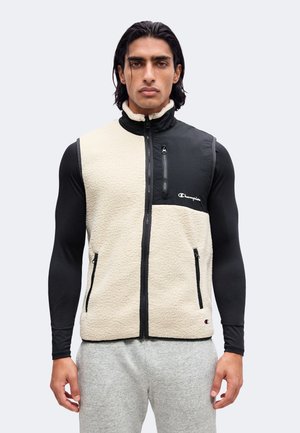 Homme portant une veste polaire zippée beige et noire par-dessus un t-shirt noir à manches longues et un pantalon de survêtement gris, faisant face à un fond uni.