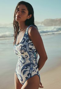 Maillot de bain une pièce avec une base blanche ornée de motifs floraux bleus et de volutes. Encolure dégagée et jambes coupées haut. Cheveux mouillés, décor de plage.