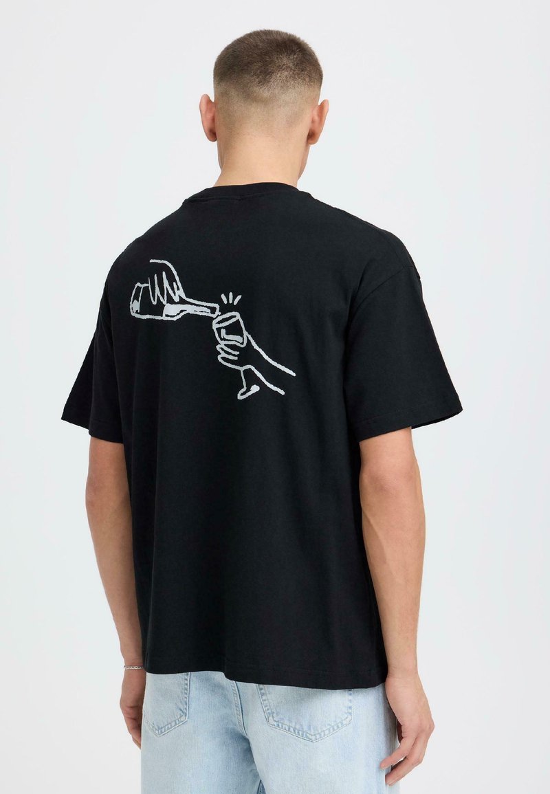 Homme aux cheveux courts portant un t-shirt noir avec un dessin au trait blanc d'une main versant une boisson dans un verre dans le dos.