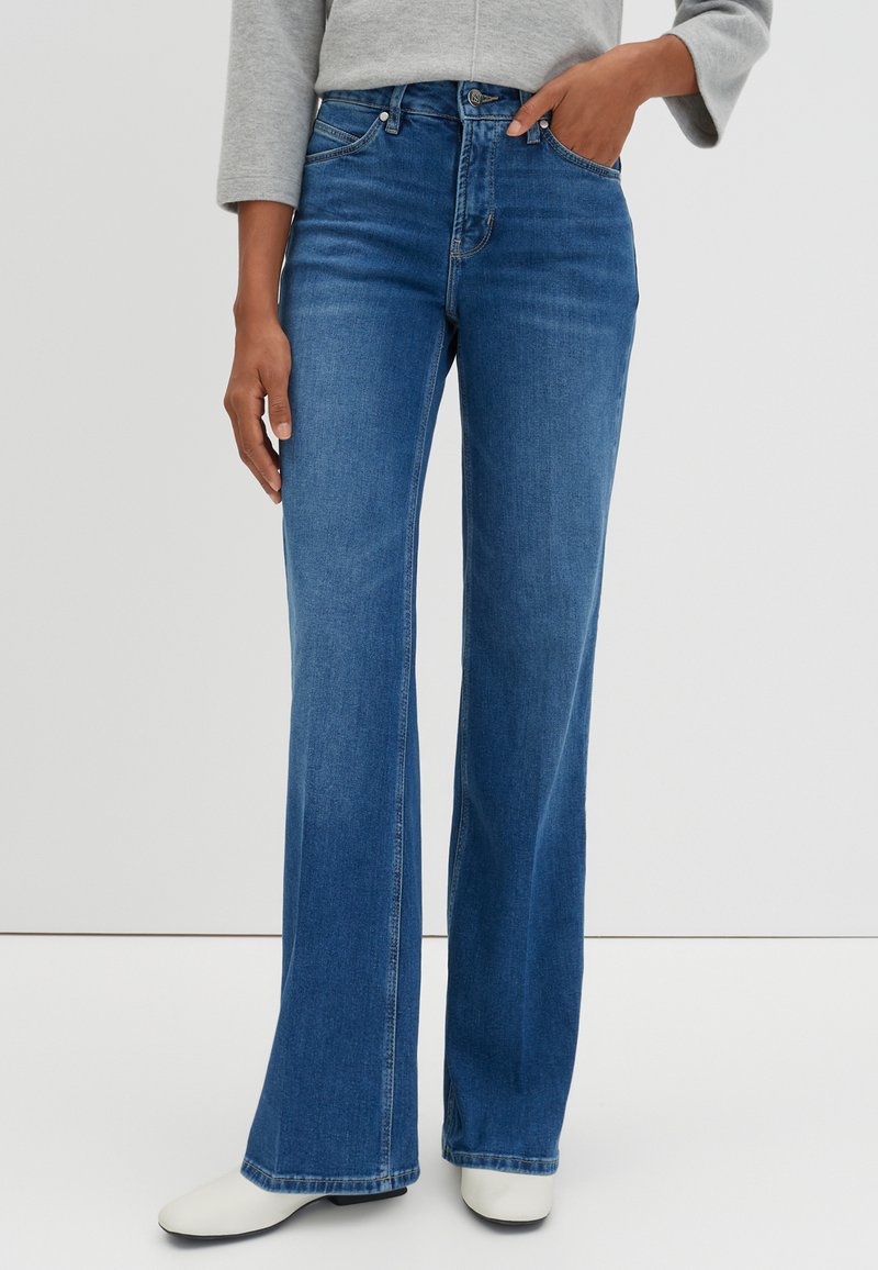 Jean flare bleu en denim, avec une taille haute, des poches avant et des coutures visibles. Assorti à des bottes blanches à bout pointu.