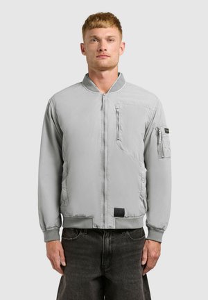 Férfi világosszürke cipzáras bomber dzsekiben, cipzáras zsebekkel a mellkasán és a bal ujján, sötét farmernadrággal párosítva.