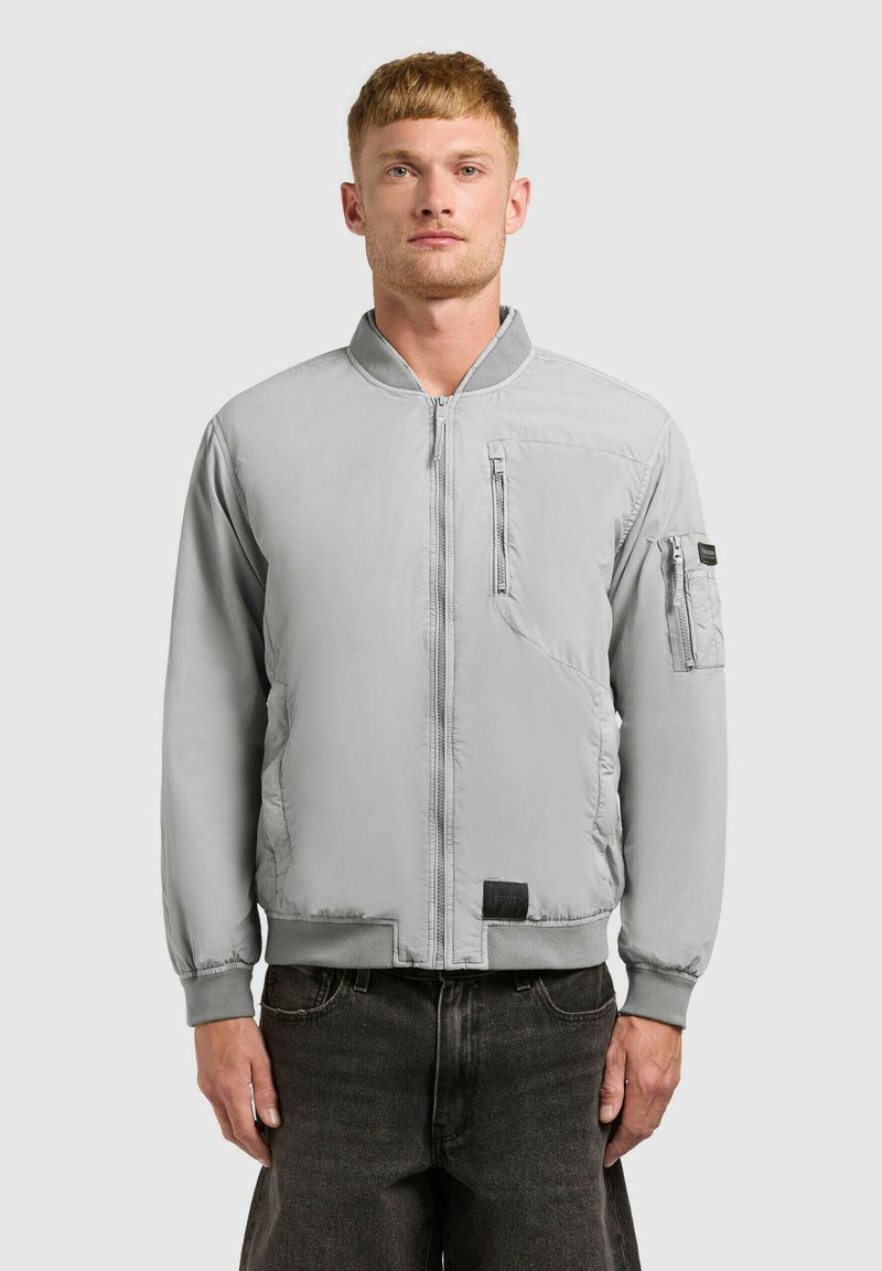 Homme portant une veste bomber légère gris clair à fermeture éclair avec poches zippées sur la poitrine et la manche gauche, associée à un jean foncé.