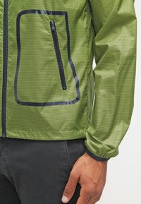 Green jacket featuring a textured honeycomb pattern, a zippered pocket on the front, and black accents along the zipper and hem.