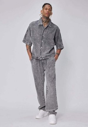 Jeune homme avec des tatouages et des dreadlocks, debout les mains dans les poches, portant une chemise ample grise délavée à manches courtes et un pantalon assorti avec des baskets blanches.