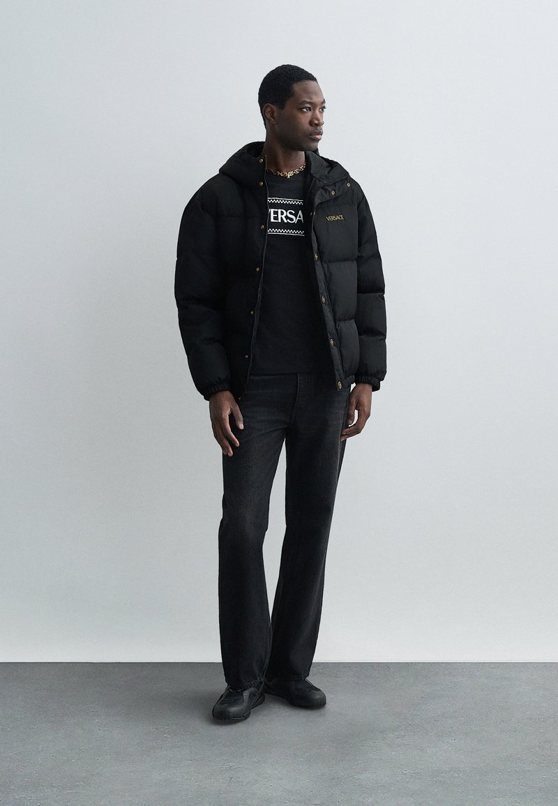 Sort pufferjakke med huva, med lynlås og lille logo; båret over en sort graphic t-shirt og mørke jeans, parret med sorte sko.