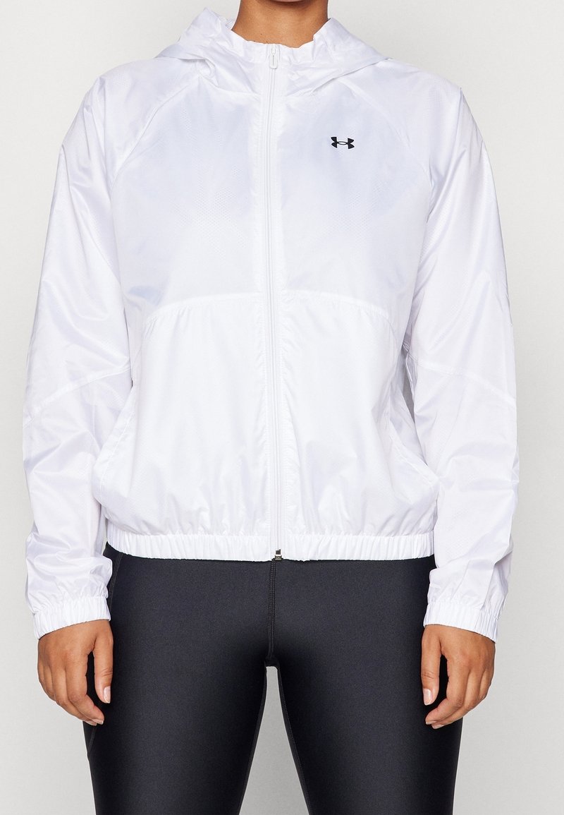 Personne portant une veste à capuche blanche zippée avec des poignets et une taille élastiques, associée à un pantalon noir ajusté.