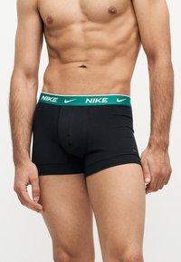 Ευχάριστο αρσενικό μοντέλο που φοράει μαύρα μποξεράκια Nike με πράσινη ζώνη και λευκά λογότυπα Nike, εμφανίζεται από τη μέση μέχρι το μέσο του μηρού.
