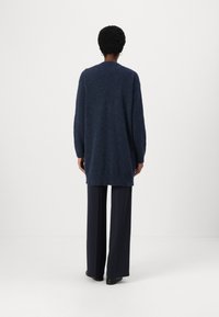Cardigan lung din navy, dintr-un material moale și pufos, cu o croială lejeră și mansete cu model în dungi. Asortat cu pantaloni largi din navy.