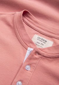 Růžová bavlněná košile s kulatým límcem, légy a třemi bílými knoflíky. Štítek uvádí "ombre bavlněná řada, velikost M, 100 % čistá bavlna."