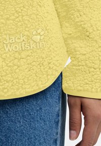 Giacca in felpa gialla con superficie testurizzata, caratterizzata da un logo con impronta di zampa. Le maniche sono morbide, i bordi sono cuciti, indossata con jeans blu.