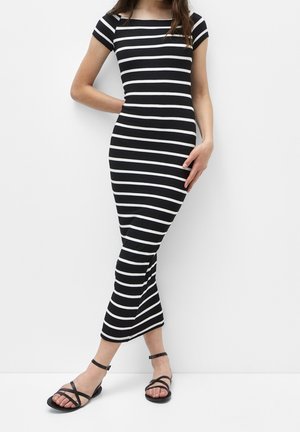 Femme portant une robe aux rayures horizontales noires et blanches, à manches courtes et longueur cheville, avec des sandales plates noires à lanières, debout contre un mur blanc.