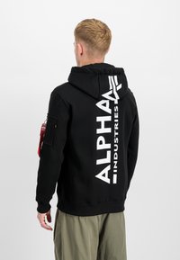 Hoodie preto com um grande logótipo branco "ALPHA INDUSTRIES" nas costas, um bolso frontal e um bolso com fecho de correr na manga, apresentando uma etiqueta vermelha.