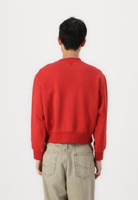 Rød sweatshirt med rund hals, ribbestrikkede ermer og nederkant. Baksiden har en V-søm detalj, båret over lyse blå denimbukser.
