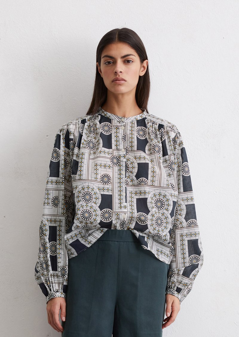 Langemouwenblouse van een geometrisch patroon in grijs, zwart en wit. Heeft geplooide schouderdetails en een hoge halslijn.